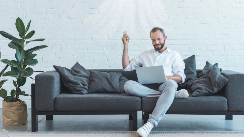 A man relaxing on a couch, using a remote to control the air conditioning in a modern living room, serviced by a trusted air conditioning company.