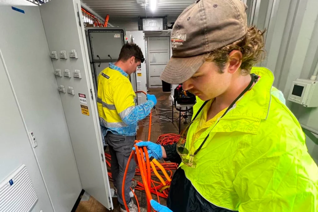 Two electricians in high-visibility jackets handle orange cables inside an industrial electrical room.