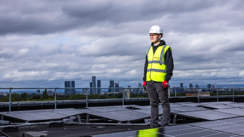 A solar technician in safety gear stands on a rooftop with solar panels to ensure the system works efficiently with a reliable solar battery.