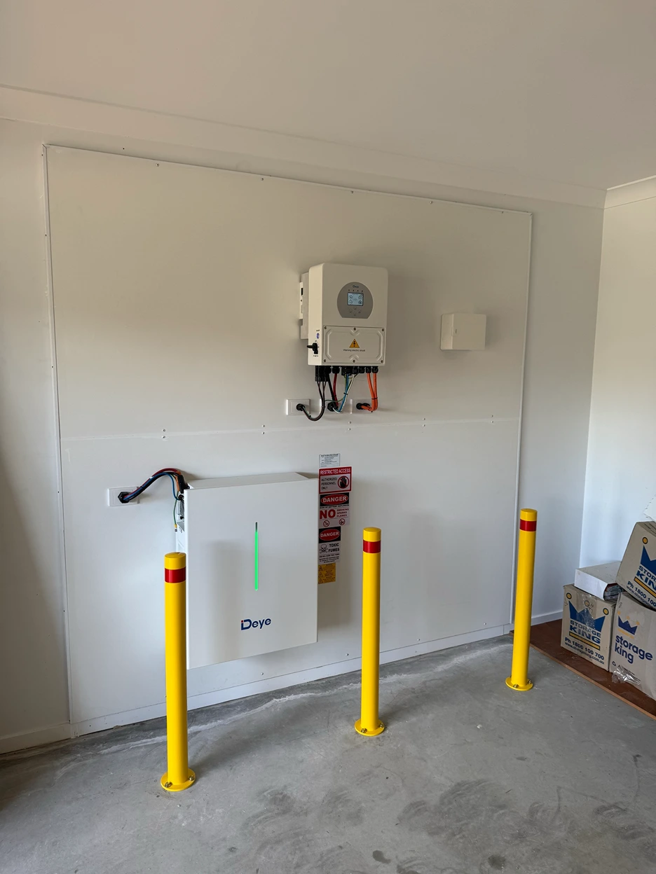 Indoor solar battery and inverter system mounted on a garage wall, protected by three yellow safety bollards.