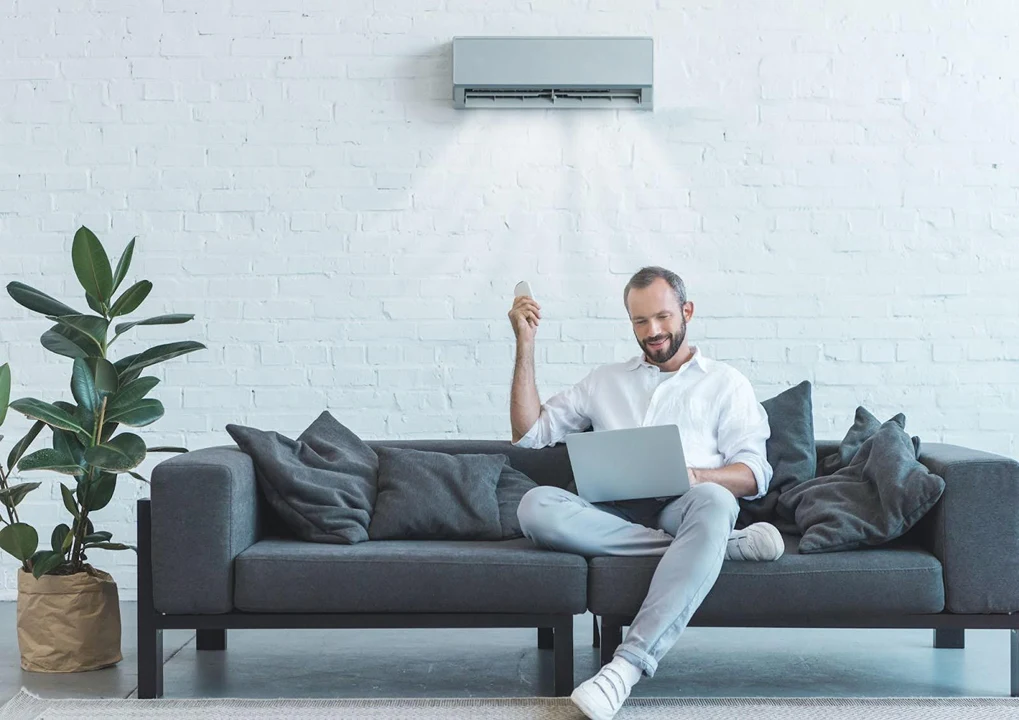 A man relaxing on a couch, using a remote to control the air conditioning in a modern living room, serviced by a trusted air conditioning company.