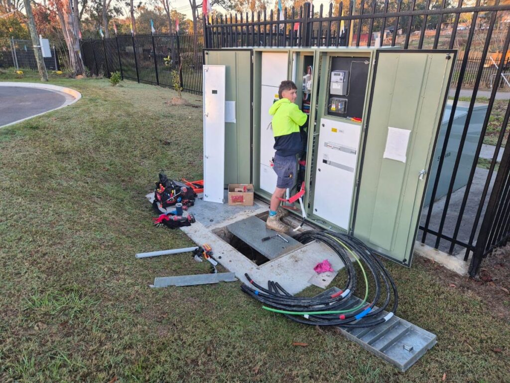 Electrician working at an outdoor pillar switchboard beside an open cable pit