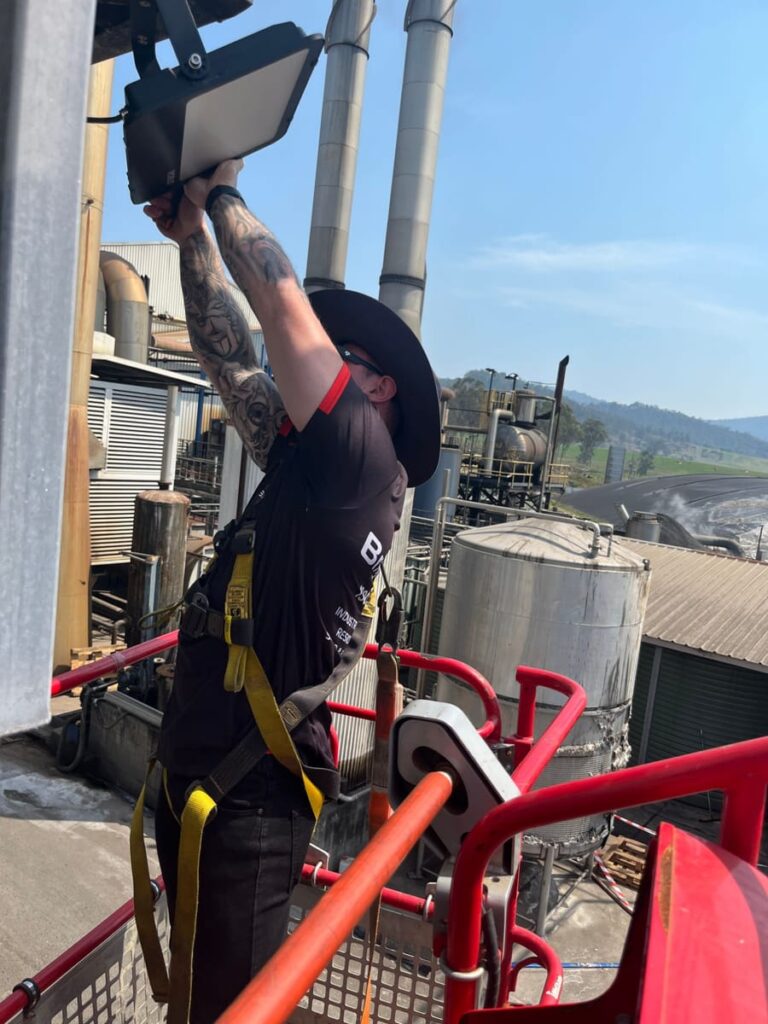 Electrician fitting an LED floodlight from an elevated work platform at an industrial site