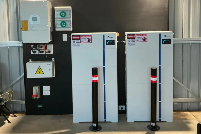 Two Deye home battery units and hybrid inverter wall-mounted in a shed with safety bollards, installed by Bright Spark Group