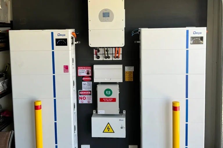 Dual Deye battery stacks with hybrid inverter and yellow safety bollards in a carport, installed by Bright Spark Group